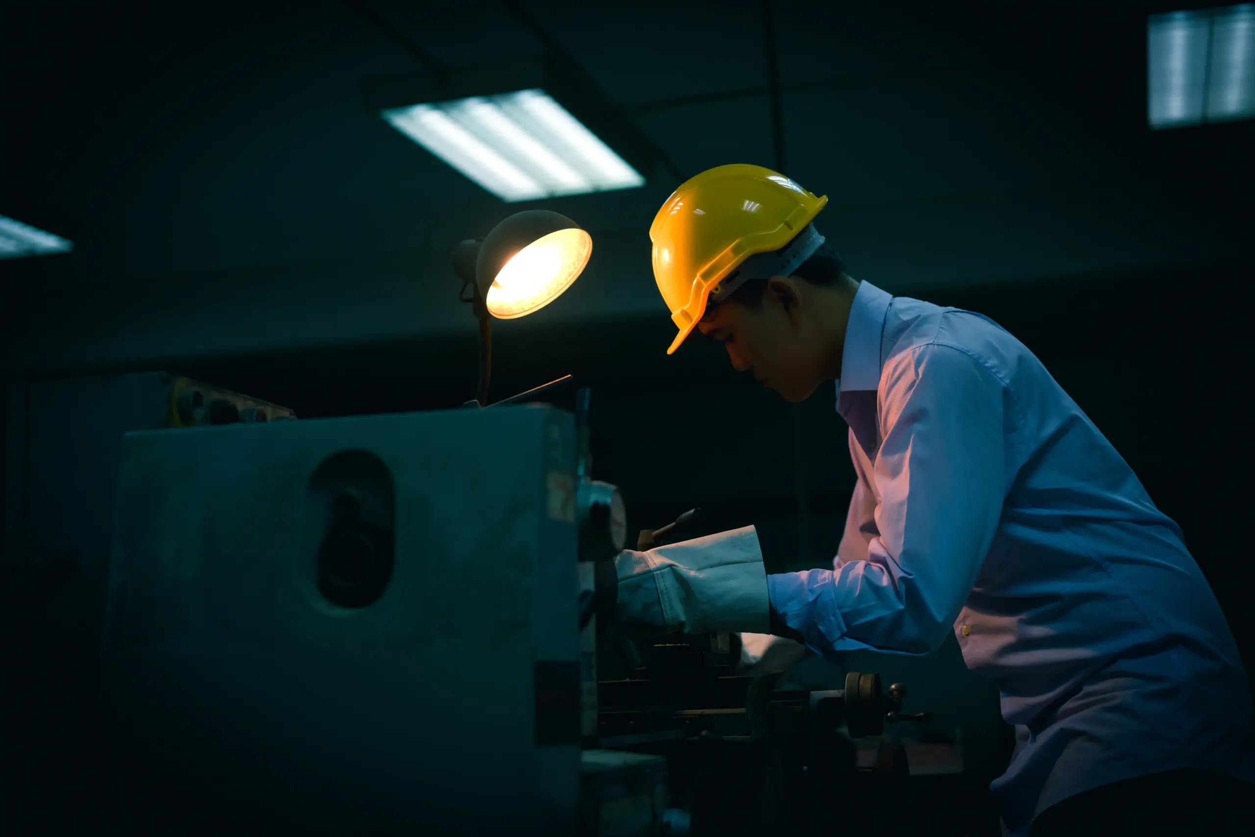 A factory worker wearing a yellow hard hat and light blue uniform leans over industrial machinery in a dimly lit manufacturing facility, with a work light illuminating the scene from above