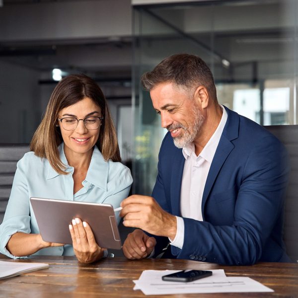 Team of diverse partners mature Latin business man and European business woman discussing project on tablet sitting at table in office. Two colleagues of professional business people working together.